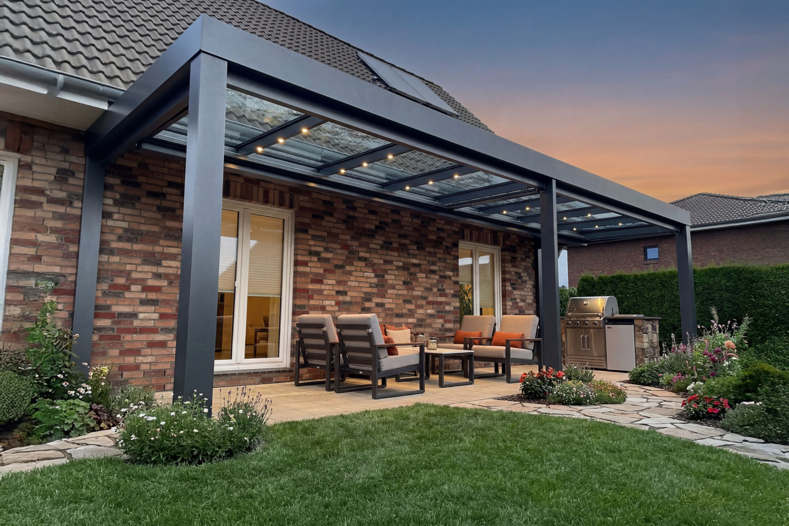 Moderne Aluminium-Terrassenüberdachung mit integrierter LED-Beleuchtung und Glasdach an einem Einfamilienhaus. Die überdachte Terrasse bietet einen stilvollen Outdoor-Wohnbereich mit Loungemöbeln und Außenküche für ganzjährigen Wetterschutz und höchsten Komfort im Garten.