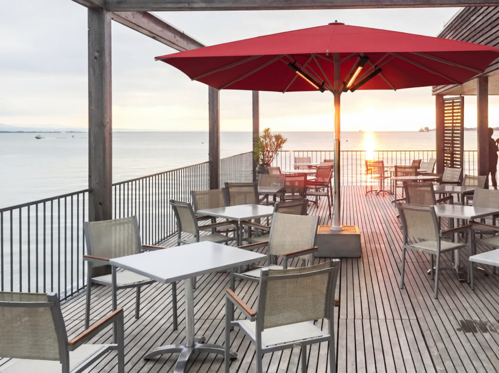 Großer roter Sonnenschirm mit integrierter Infrarotheizung auf einer modernen Restaurantterrasse direkt am Meer mit Tischen, Stühlen und Blick auf den Sonnenuntergang.