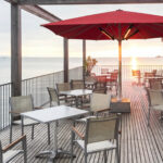 Großer roter Sonnenschirm mit integrierter Infrarotheizung auf einer modernen Restaurantterrasse direkt am Meer mit Tischen, Stühlen und Blick auf den Sonnenuntergang.