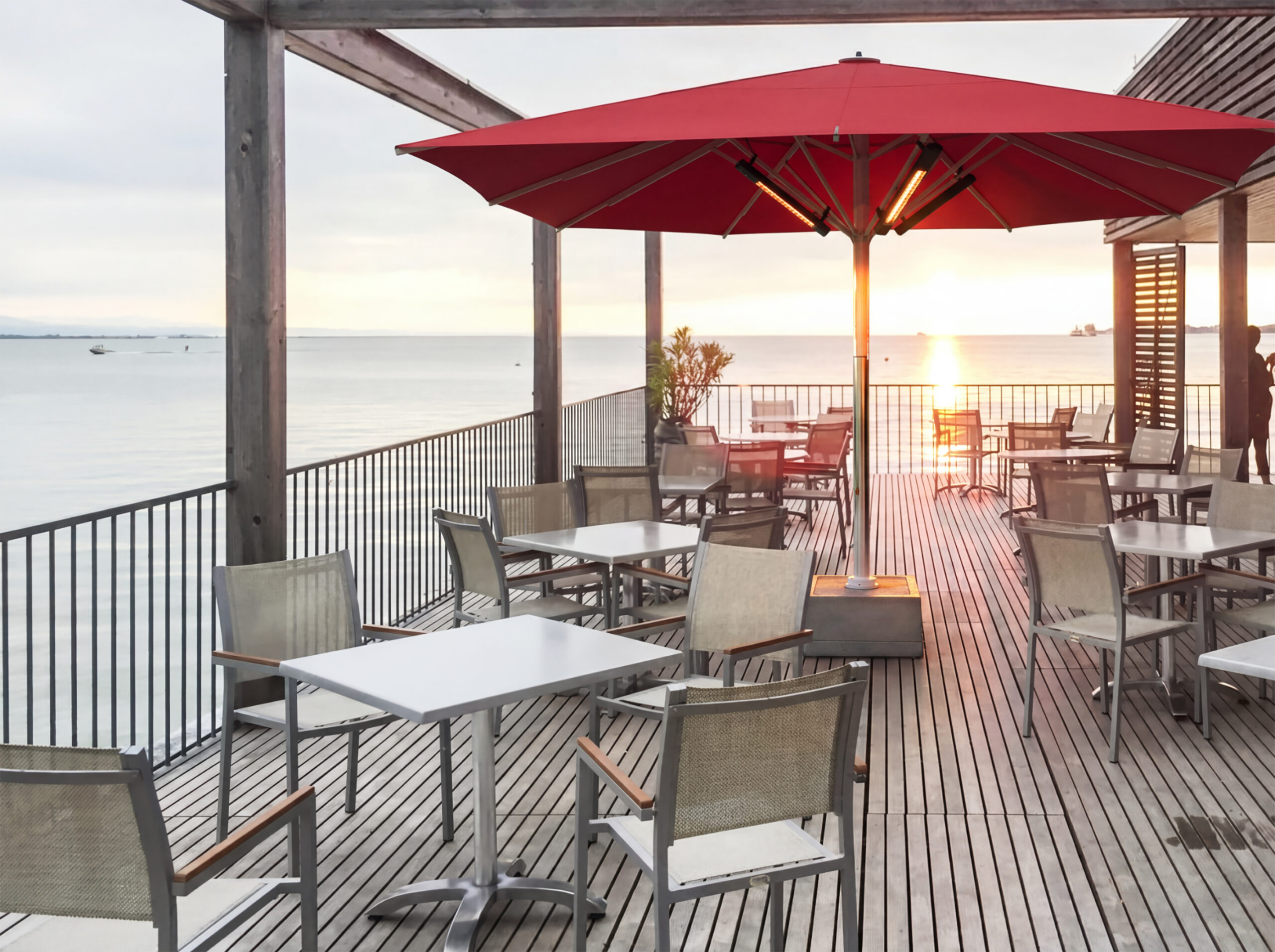 Großer roter Sonnenschirm mit integrierter Infrarotheizung auf einer modernen Restaurantterrasse direkt am Meer mit Tischen, Stühlen und Blick auf den Sonnenuntergang.