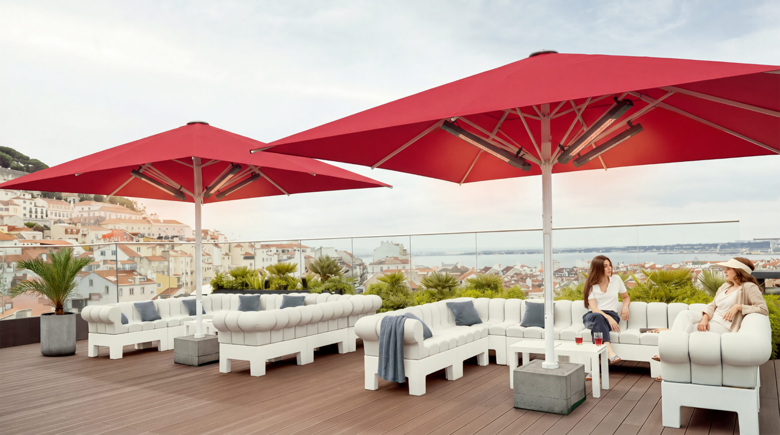 Zwei große rote Sonnenschirme auf einer modernen Dachterrasse mit weißen Loungemöbeln, zwei sitzenden Personen und Panoramablick über eine mediterrane Küstenstadt.
